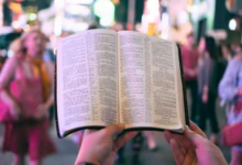 person holding bible on road with people walking on sidewalk beside buildings during nighttime